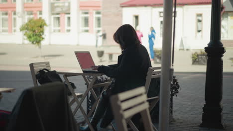 freelancer seated at outdoor cafe table working on laptop, surrounded by wooden chairs, potted plants, and a paved walkway, with blurred background of people standing