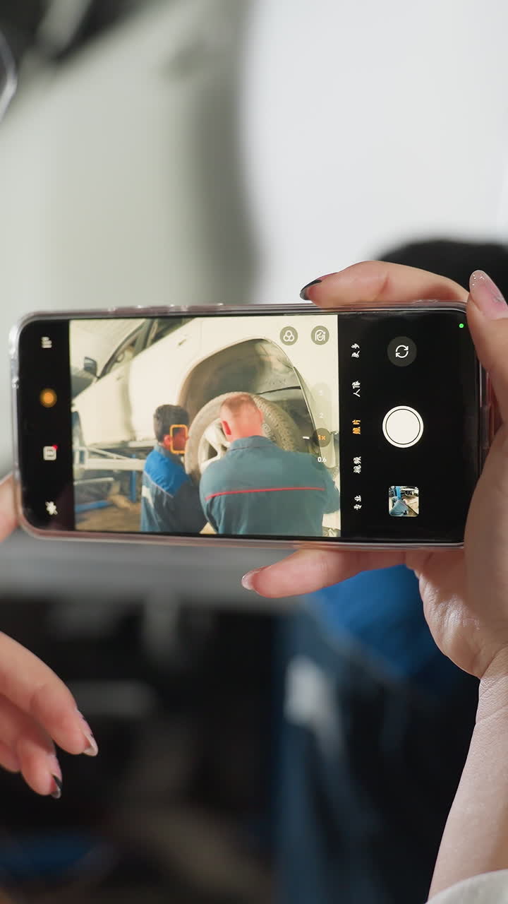 vista posterior de cerca de una mujer con bata de laboratorio usando un teléfono inteligente para tomar una foto de un mecánico llevando y arreglando neumáticos en un coche en un taller de mecánicos