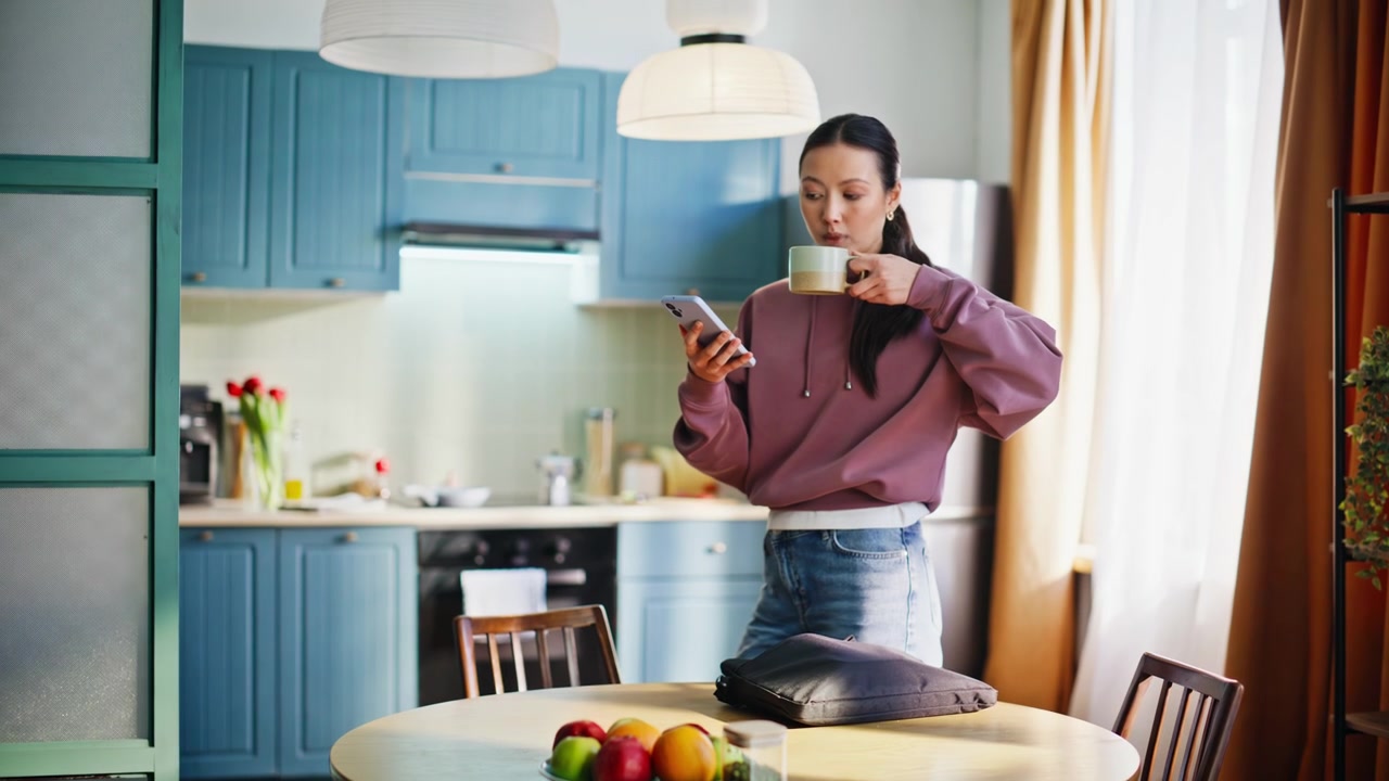 Donna asiatica che prepara la borsa del laptop, beve caffè e controlla il telefono nella cucina di casa