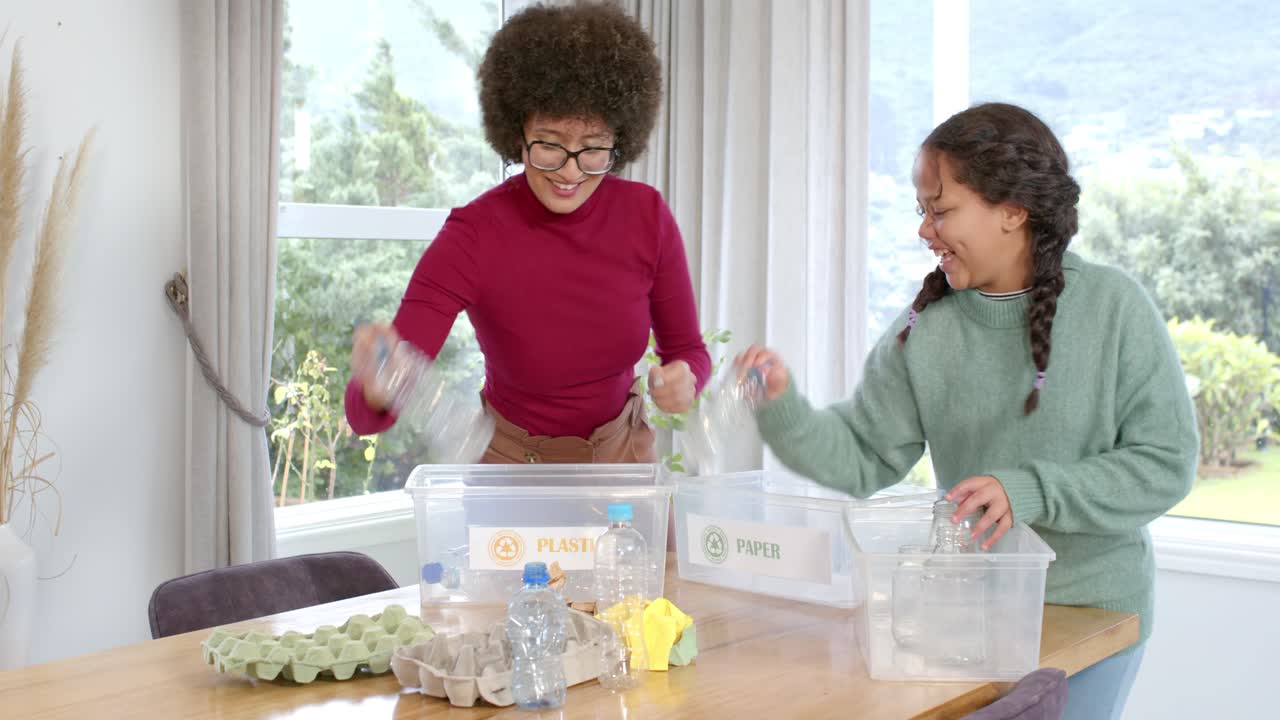 African American mom and daughter grabbing recyclables sorting into labeled bins at wooden table