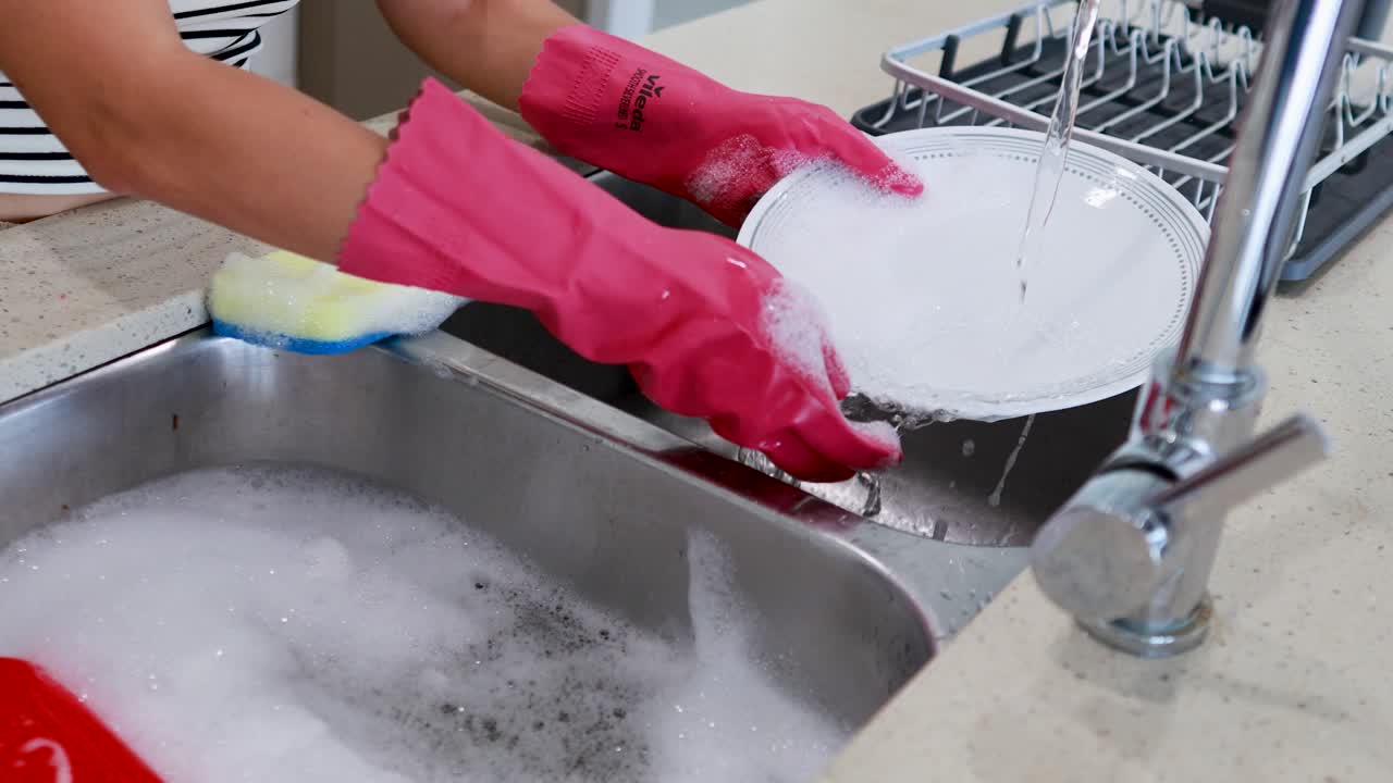 Person wearing gloves washes dishes in a bright kitchen sink. Running water and organized setup create a clean, efficient atmosphere
