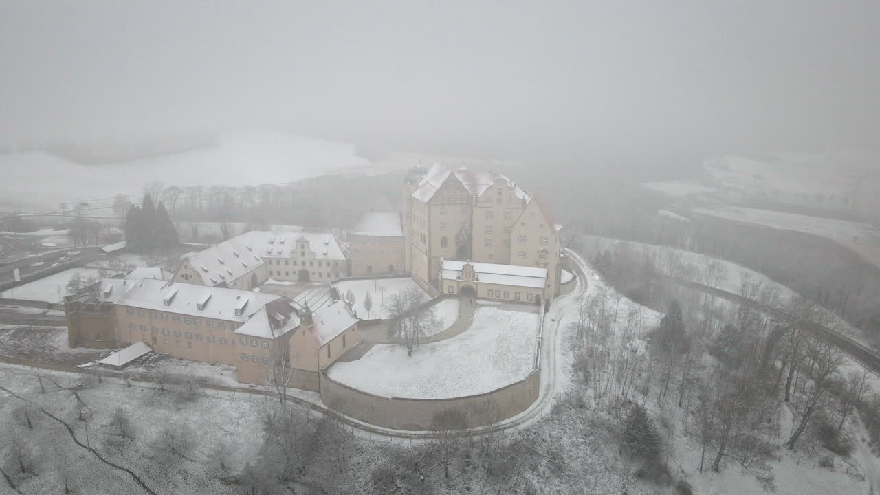 Drone pans right across Schloss Kapfenburg highlighting the castle's gabled roofs, bastion towers, and snow-laden courtyards enveloped in thick fog and white expanses. Mystical fortress reveal