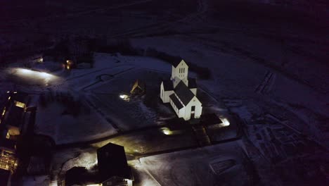 a cinematic aerial drone shot capturing a peaceful icelandic church at night, surrounded by quiet rural land and softly lit buildings beneath the moody northern sky.