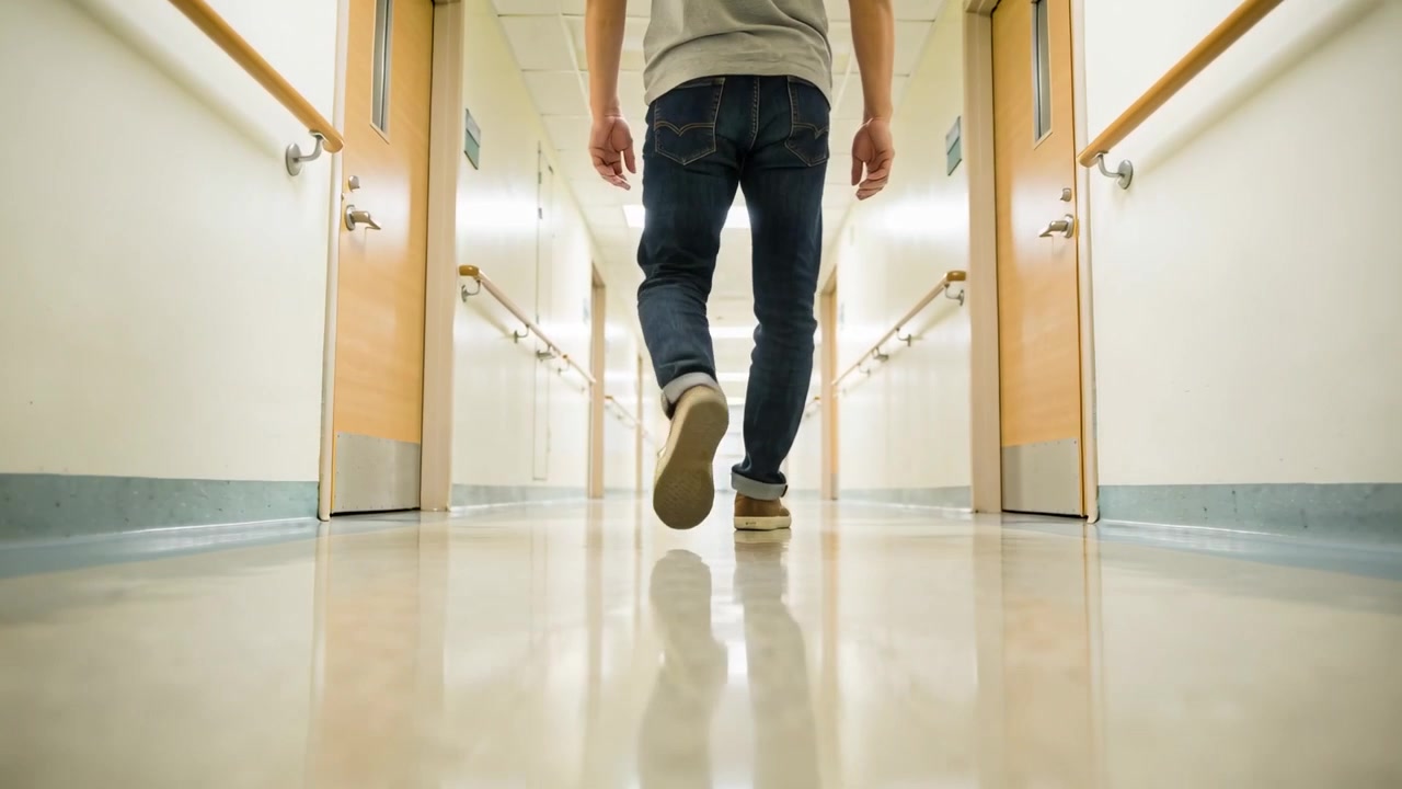 Un uomo adulto in jeans e scarpe da ginnastica che cammina verso la fine del corridoio, passando accanto a corrimano e a una zona illuminata.