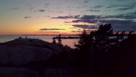 fly past inukshuk on rocky pine tree island just after sunset, drone aerial wide dolly in