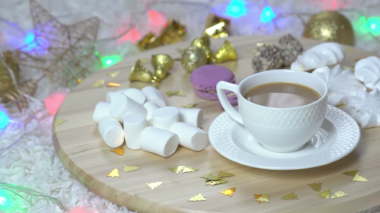 una taza de café con leche y marshmallows en salver de madera rotación de 360 grados, otros dulces y decoraciones de navidad alrededor.
