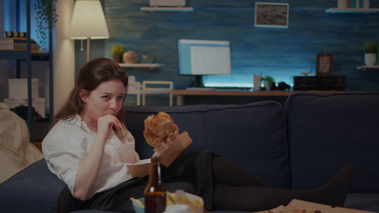 Young woman laying on couch eating burger and laughing