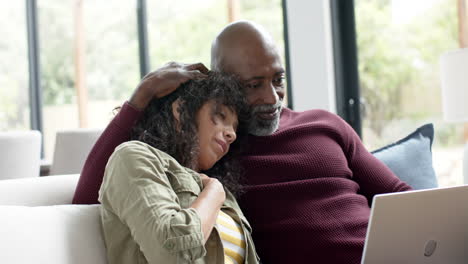 Happy-biracial-couple-sitting-on-couch-and-using-laptop-at-home,-slow-motion