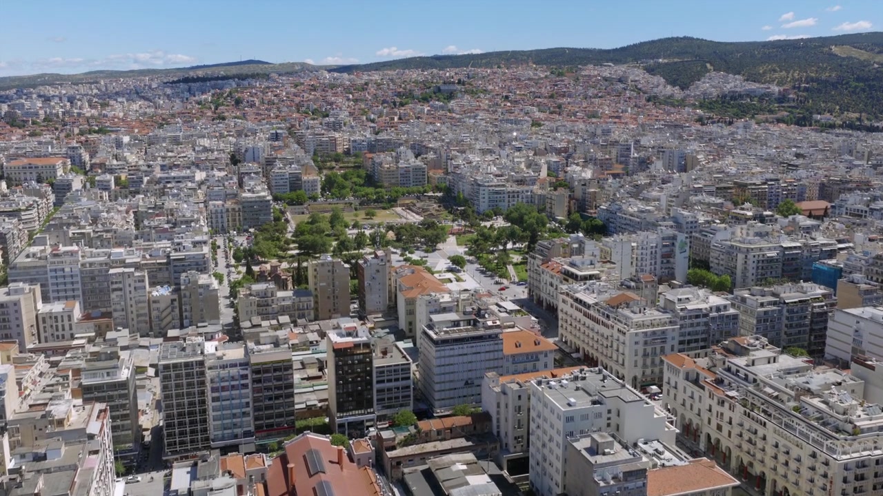 Vista aerea ravvicinata degli edifici e dei tetti del centro di Salonicco, con parchi verdi e una fitta architettura che si estende sullo sfondo.