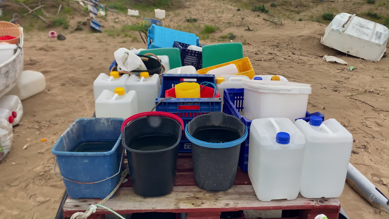 Contenitori di plastica, secchi, cassette e taniche piene d'acqua posizionate su un pallet di legno su una spiaggia sabbiosa di Sanlúcar de Barrameda.