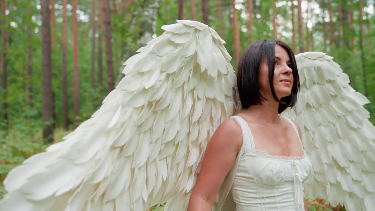 White Fairy woman with large angel wings walks gracefully through green forest, holding wooden stick, observing nature around her in serene atmosphere filled with sunlight