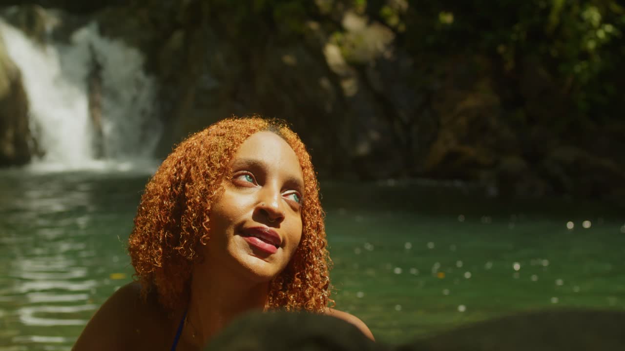 Tons de jungle vert profond entourant une fille aux cheveux roux en bikini bleu, posant près d'une cascade impétueuse.