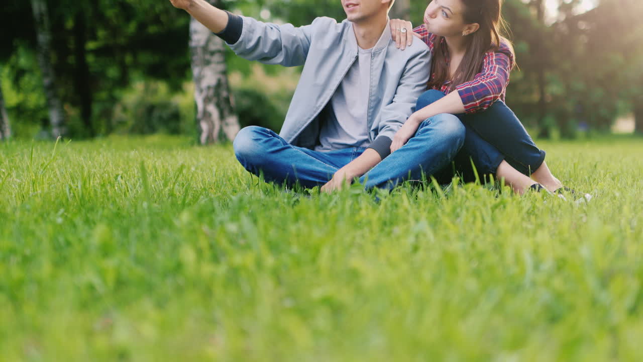그라에 공원에 앉아 재미있는 얼굴을 만드는 전화에 자신을 촬영하는 젊은 부부