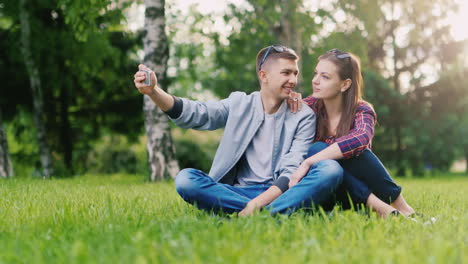 Junges-Paar-Fotografiert-Sich-Am-Telefon-Und-Macht-Lustige-Grimassen,-Die-Im-Park-Auf-Dem-Gras-Sitzen