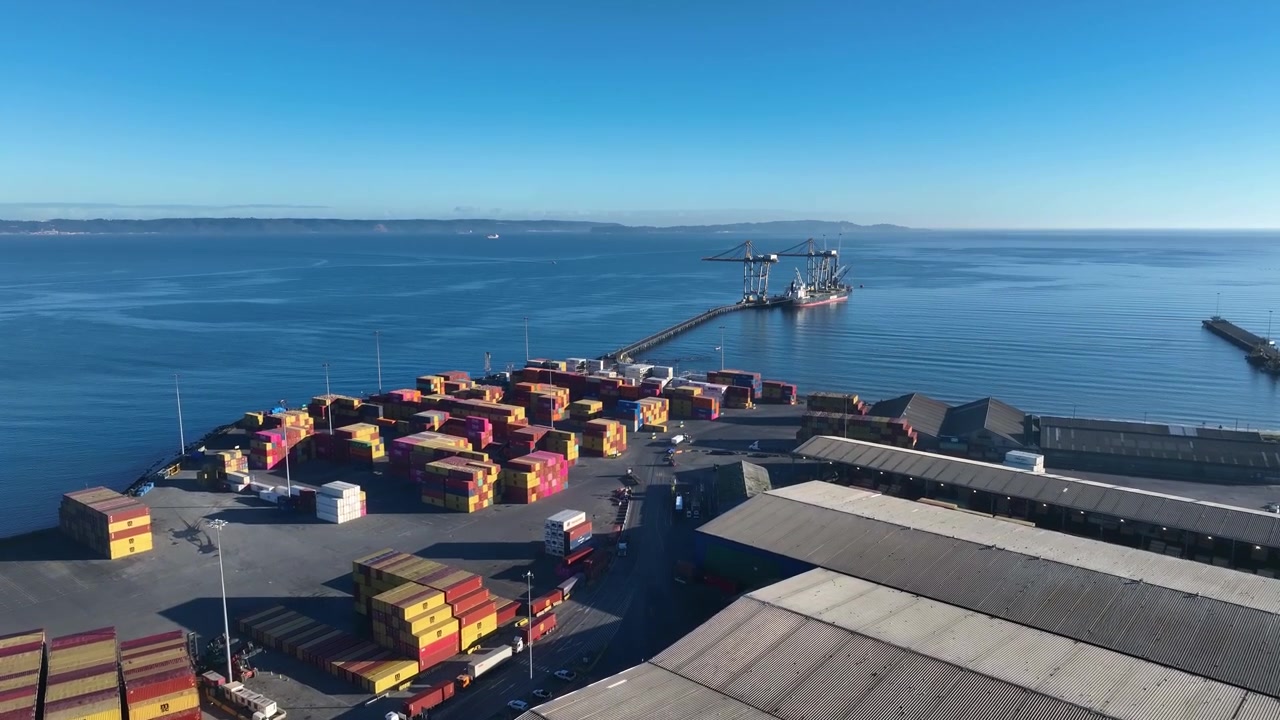 Advancing flight over stacked shipping containers and warehouses toward a loading pier in a calm coastal bay