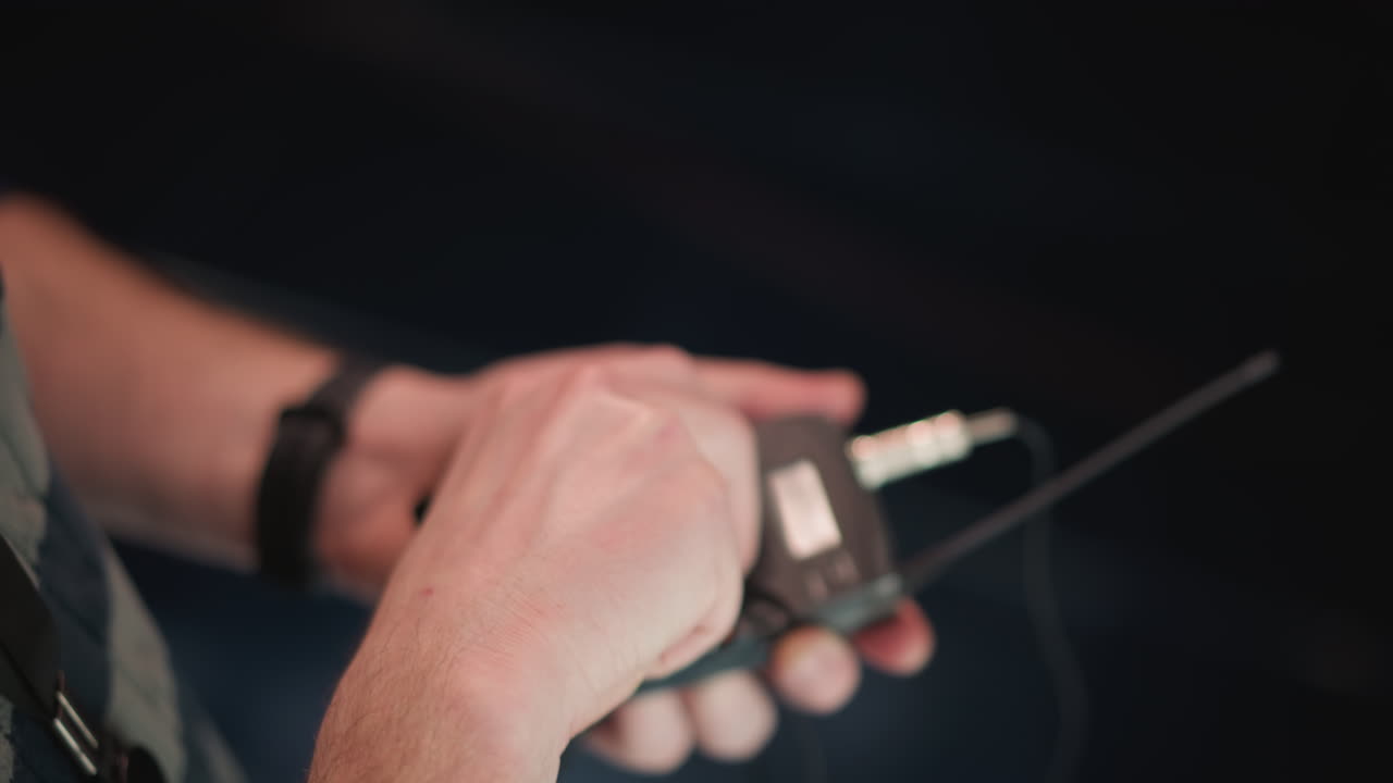 hands male technician wearing watch attaches cable to wireless microphone transmitter unit while inspecting batteries under bright studio lighting grid rigging visible behind, conveying behind scenes