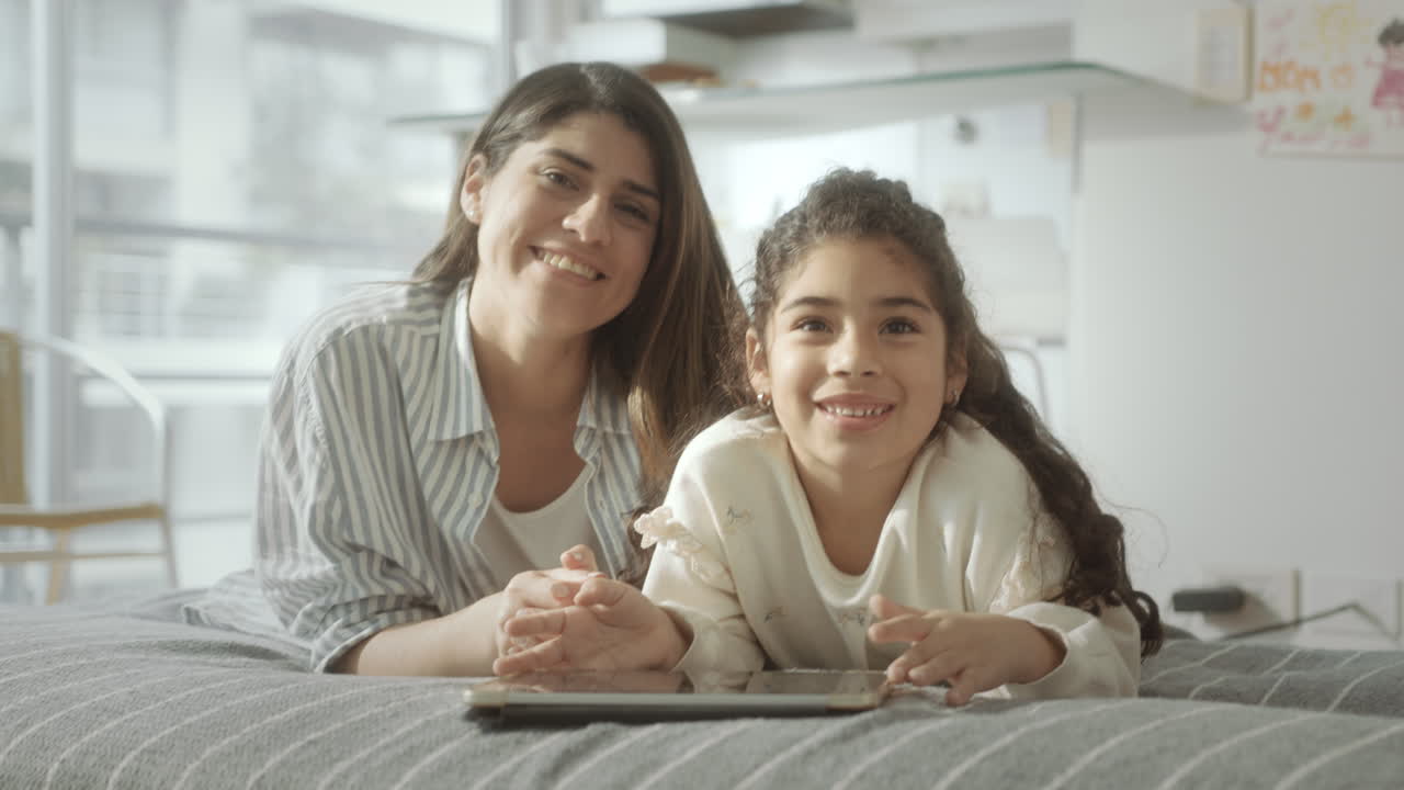 Retrato de madre feliz y su hija pequeña con tableta digital en la cama