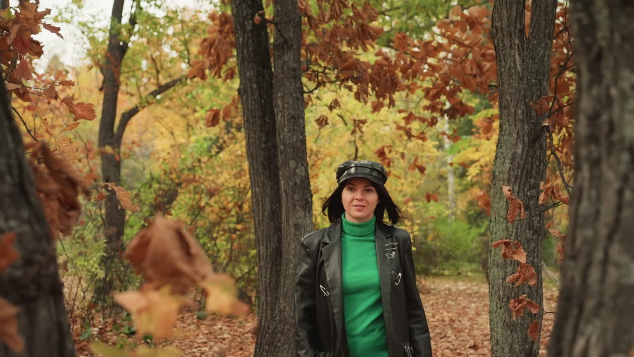 caucasian woman in leather jacket walks between oak trees, autumn leaves falling, contemplative expression, slow camera pull, serene forest path, emerald sweater contrast, overcast soft light, rustic