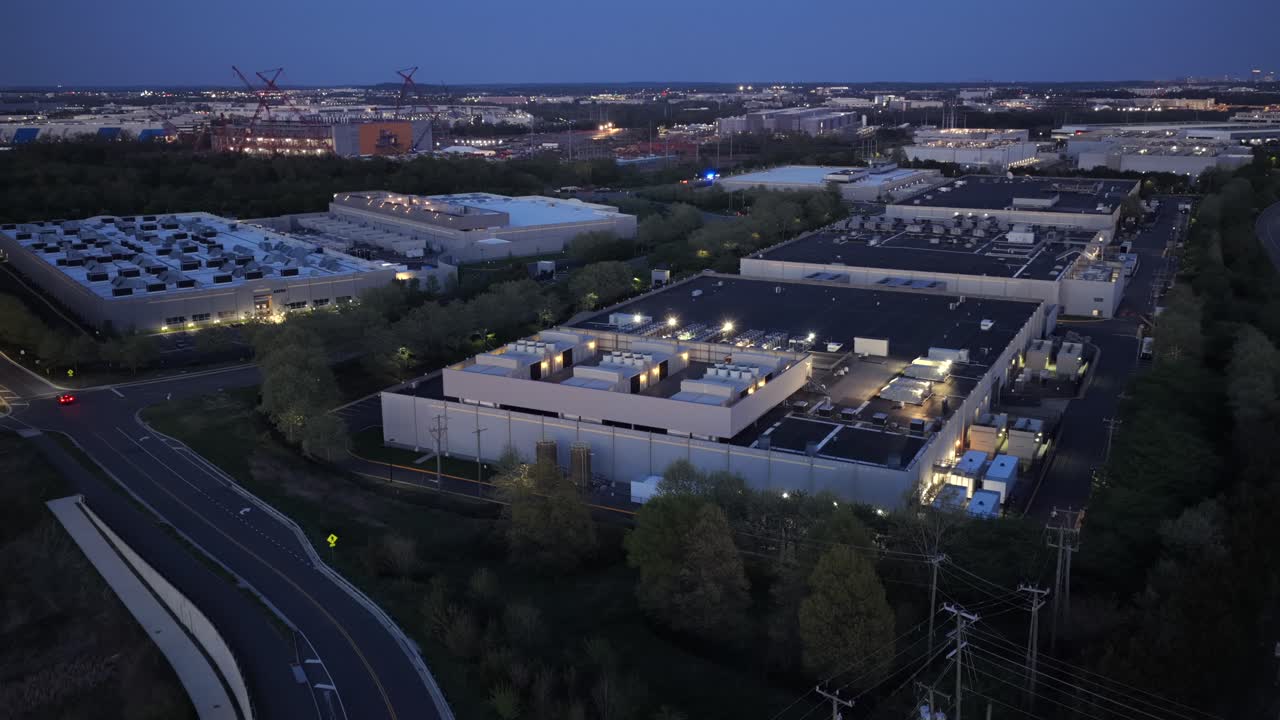 Veduta aerea al crepuscolo di un campus di data center illuminato ad Ashburn, Virginia, con strade, auto di passaggio e moderne strutture server sotto il cielo dell'ora blu nel corridoio tecnologico degli Stati Uniti. Inquadratura ampia.