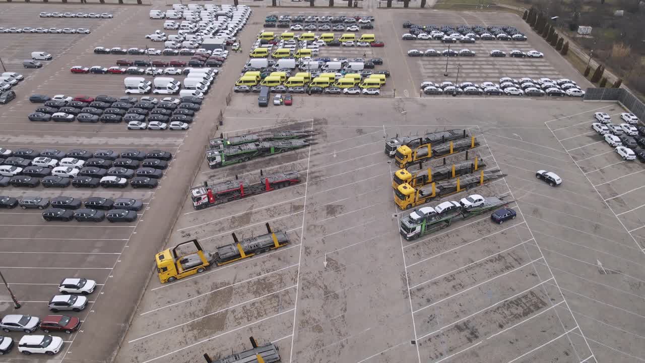 An aerial video of a car depot showcasing rows of parked cars, vans, and yellow commercial vehicles.