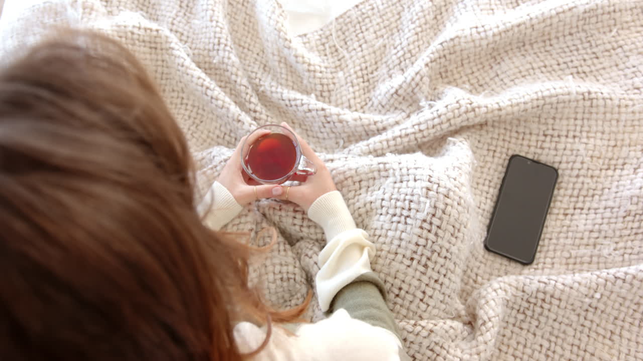 Holding cup of tea, woman relaxing on bed with smartphone nearby