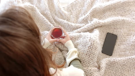 holding cup of tea, woman relaxing on bed with smartphone nearby