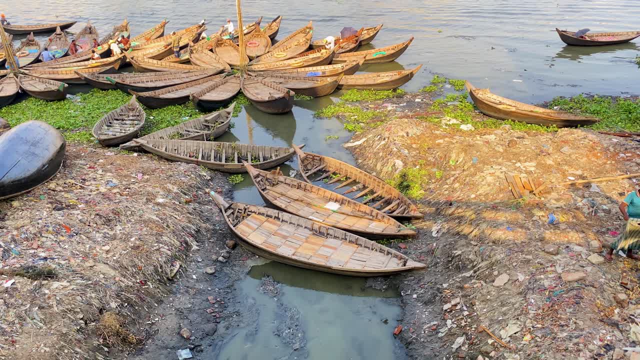 Wooden boats docked on polluted river in Dhaka, Bangladesh with city buildings in the background. Represents water pollution and river pollution.