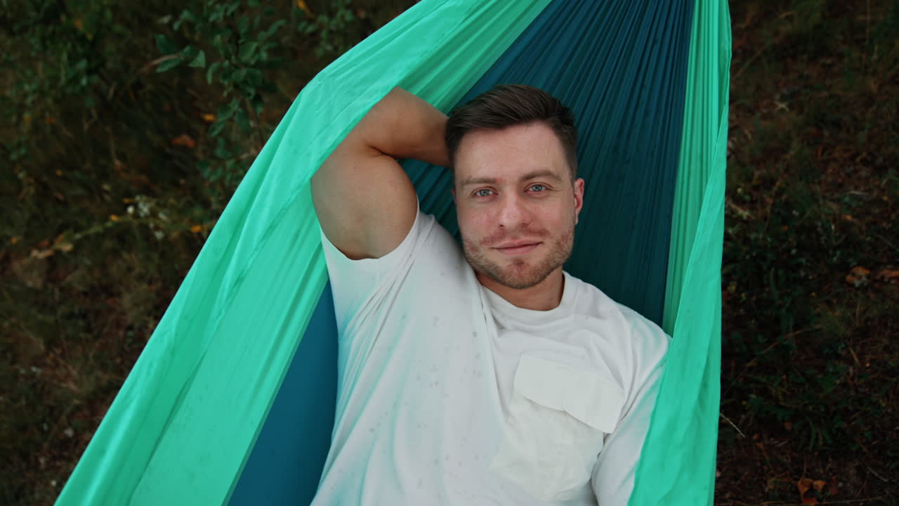 Smiling relaxed Caucasian man lies comfortably in a hammock. Man having vacation in nature. Top view.