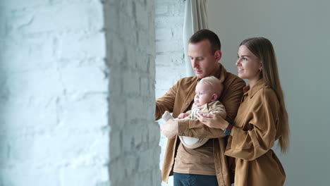 husband and wife stand at the window with their infant son. dad calms down crying baby showing something in the window.
