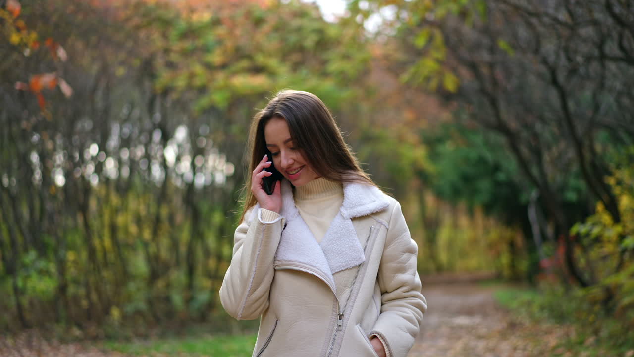 Beautiful dark-haired lady in white jacket walking slowly by the autumn wood. Attractive woman speaks on the phone continuing her walk.