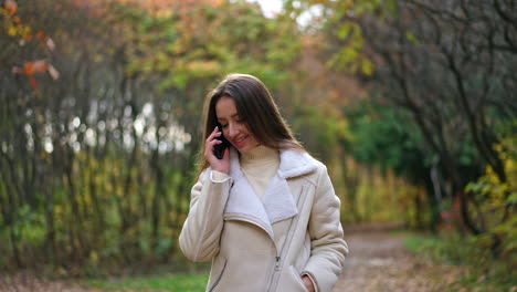 beautiful dark-haired lady in white jacket walking slowly by the autumn wood. attractive woman speaks on the phone continuing her walk.