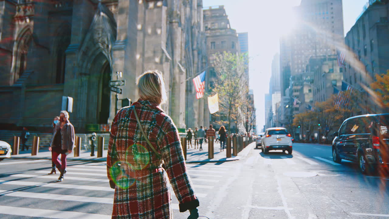 Nueva York, EE. UU., 5 de octubre de 2025: Mujer con abrigo de cuadros cruzando la calle en St. Patrick's. Mujer caminando por un paso de peatones con la Catedral de San Patricio y un fuerte destello solar de fondo