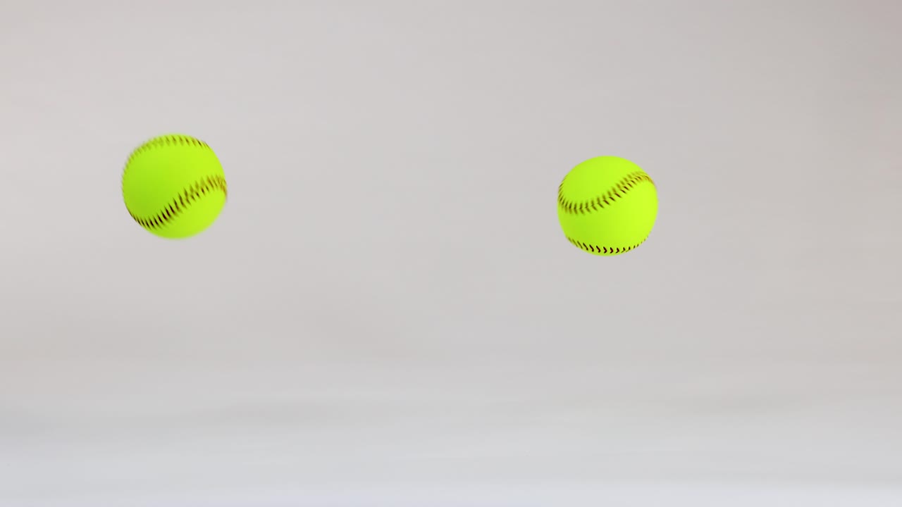 Two optic yellow softballs bounce and roll across a clean white studio floor background