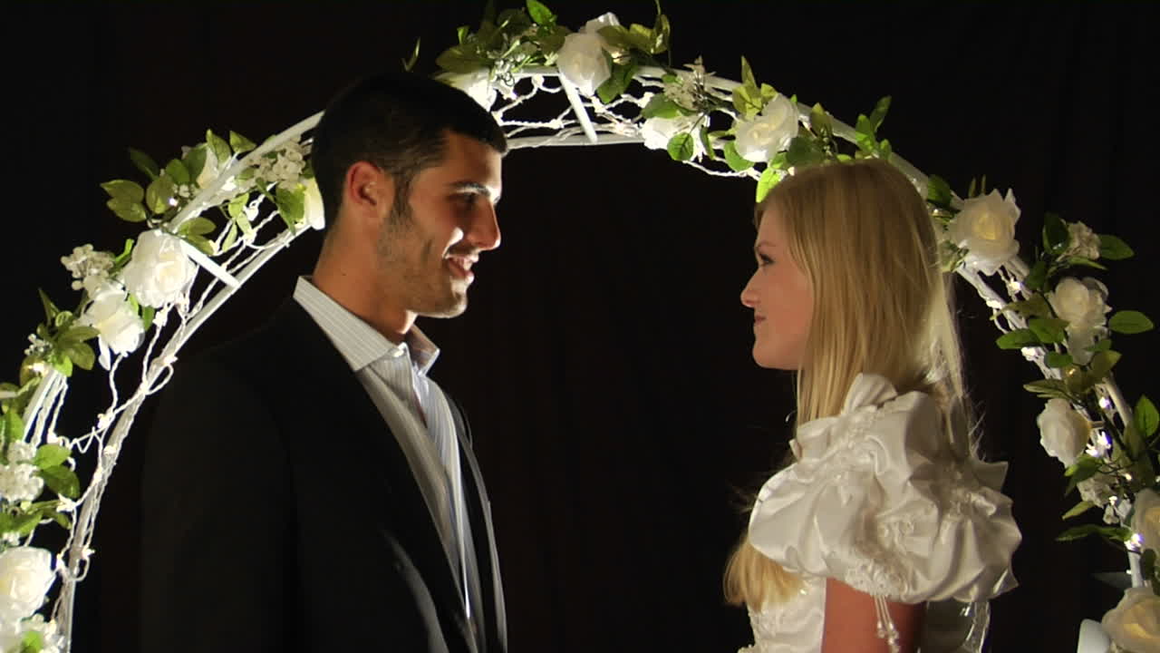 la novia y el novio se paran bajo un arco de flores mientras el hombre desliza un anillo en el dedo de la mujer