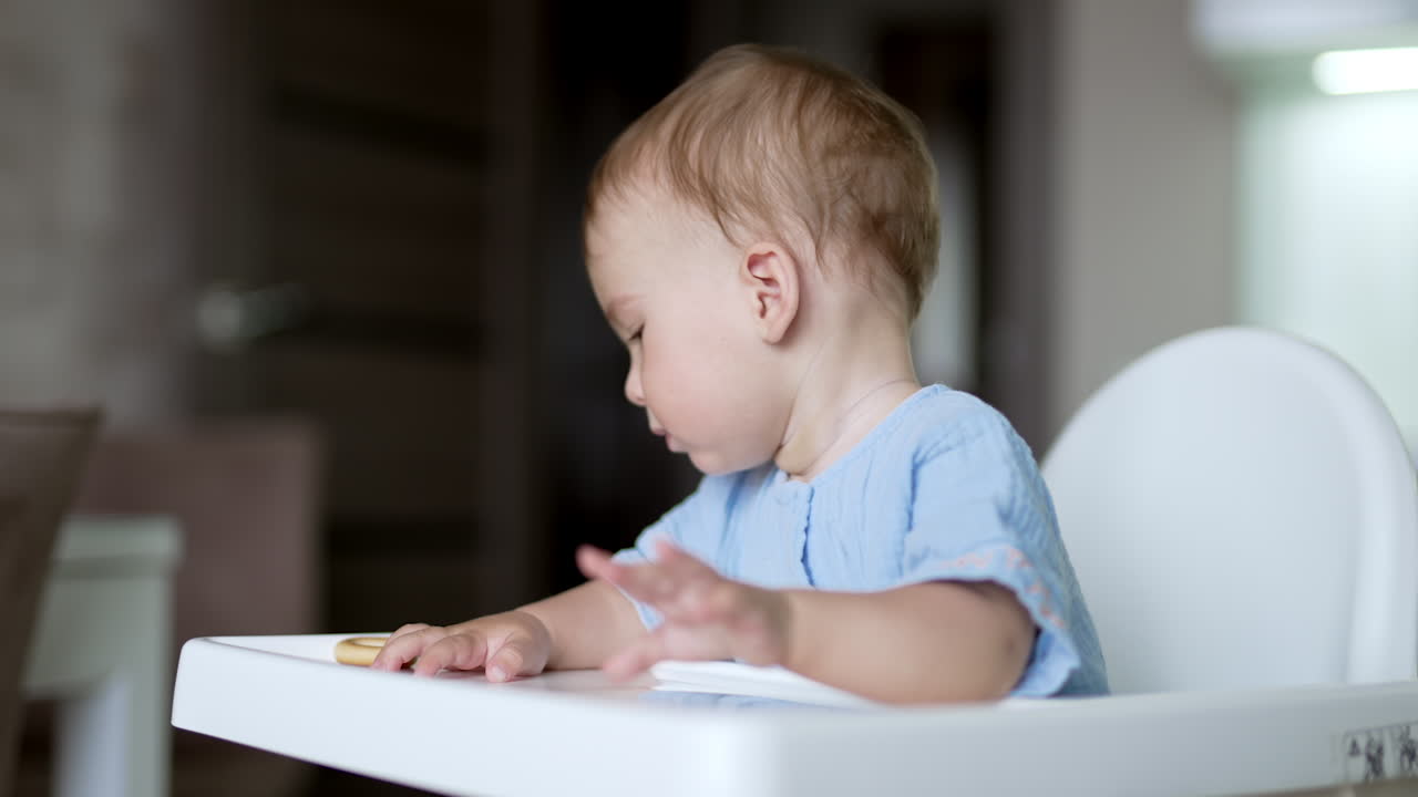 Lovely Caucasian child at the feeding table eating a bagel. Cute boy playing with food. Close up. Blurred backdrop.