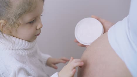 little girl applies cream on pregnant mother belly in room