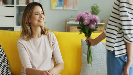 Süßes-Blondes-Jugendlich-Mädchen,-Das-Blumen-über-Dem-Rücken-Versteckt-Und-Es-Ihrer-Mutter-Im-Wohnzimmer-Gibt