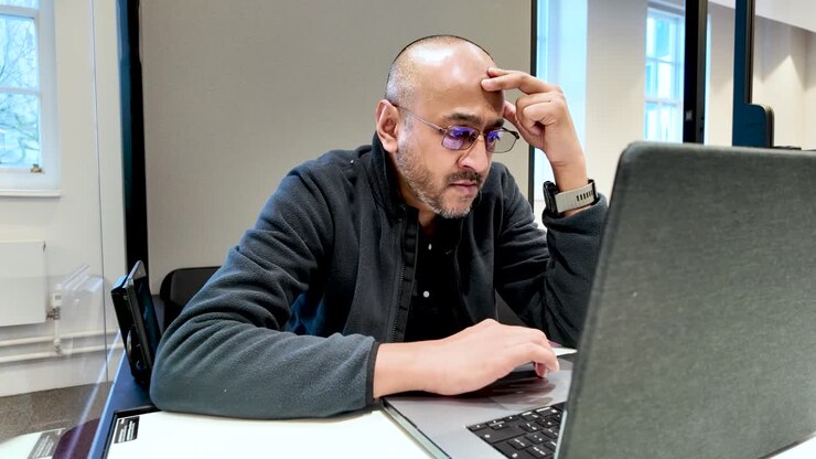 South asian male professional concentrating on his laptop, hand on forehead, in a contemporary office pod