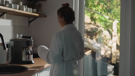 woman makes coffee with machine in kitchen. relaxed lady in terry bathrobe comes to coffee maker preparing hot aromatic drink at home. happy morning