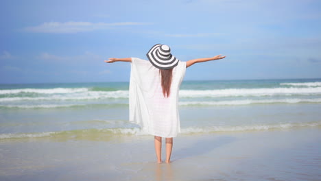 back of joyful woman in white tunic on sandy beach raising arms in front of sea waves and horizon