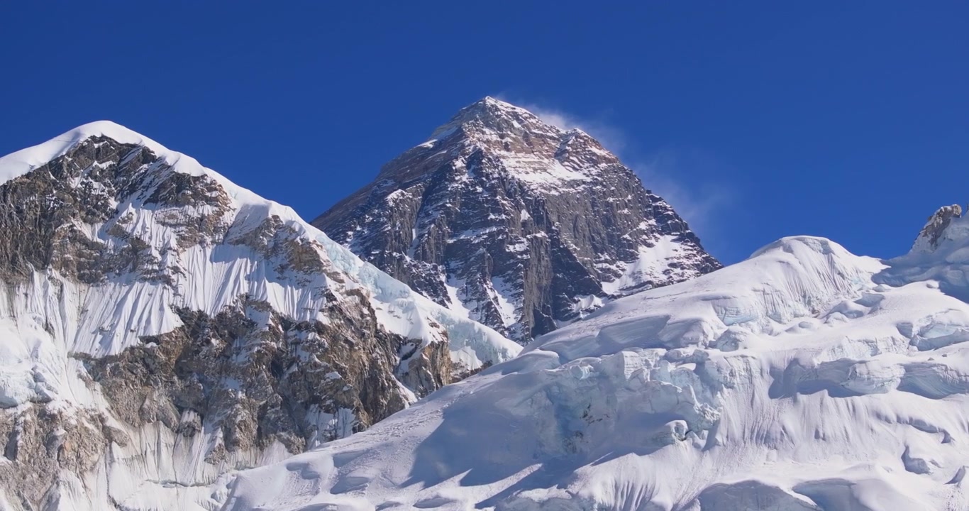 Regione dell'Everest, Nepal, ripresa aerea laterale con drone che cattura la scura cima rocciosa del Monte Everest con un pennacchio di neve che si solleva dalla vetta dietro i pendii innevati del Nuptse