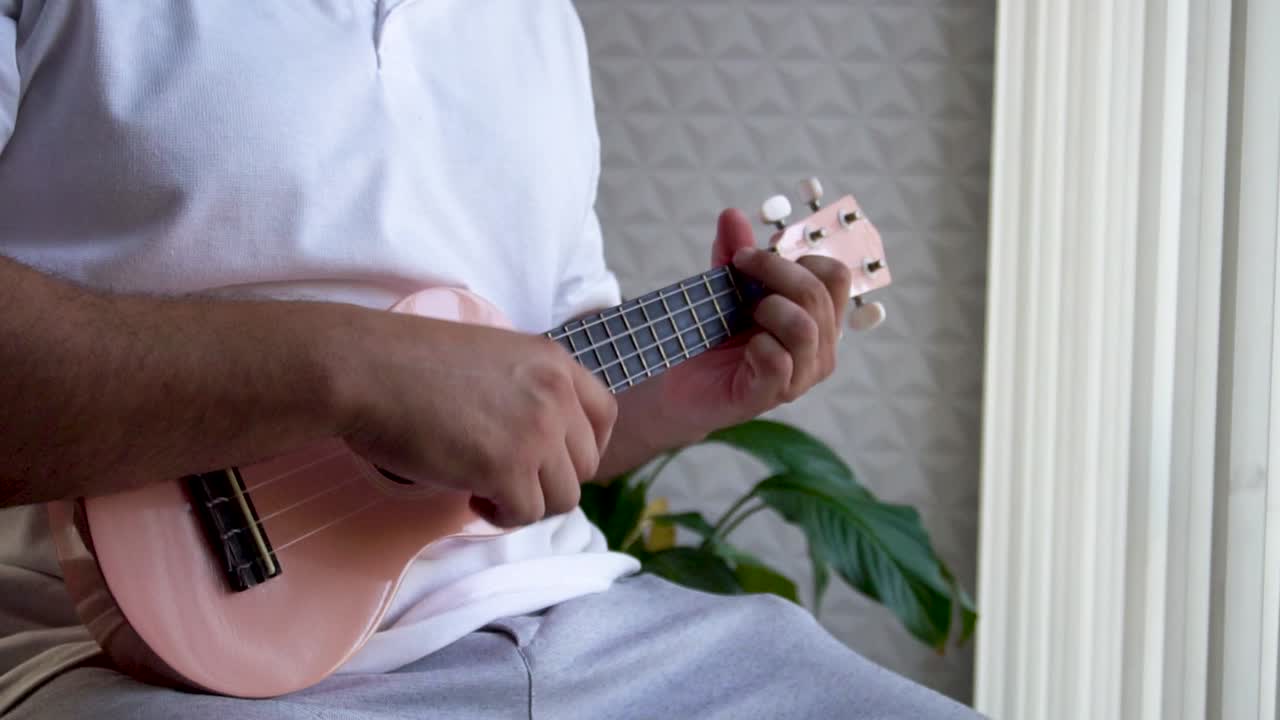 hombre de mediana edad 30 y con cuentas está jugando y silbando con su ukulele rosado al lado de una gran ventana y cerca de una planta verde