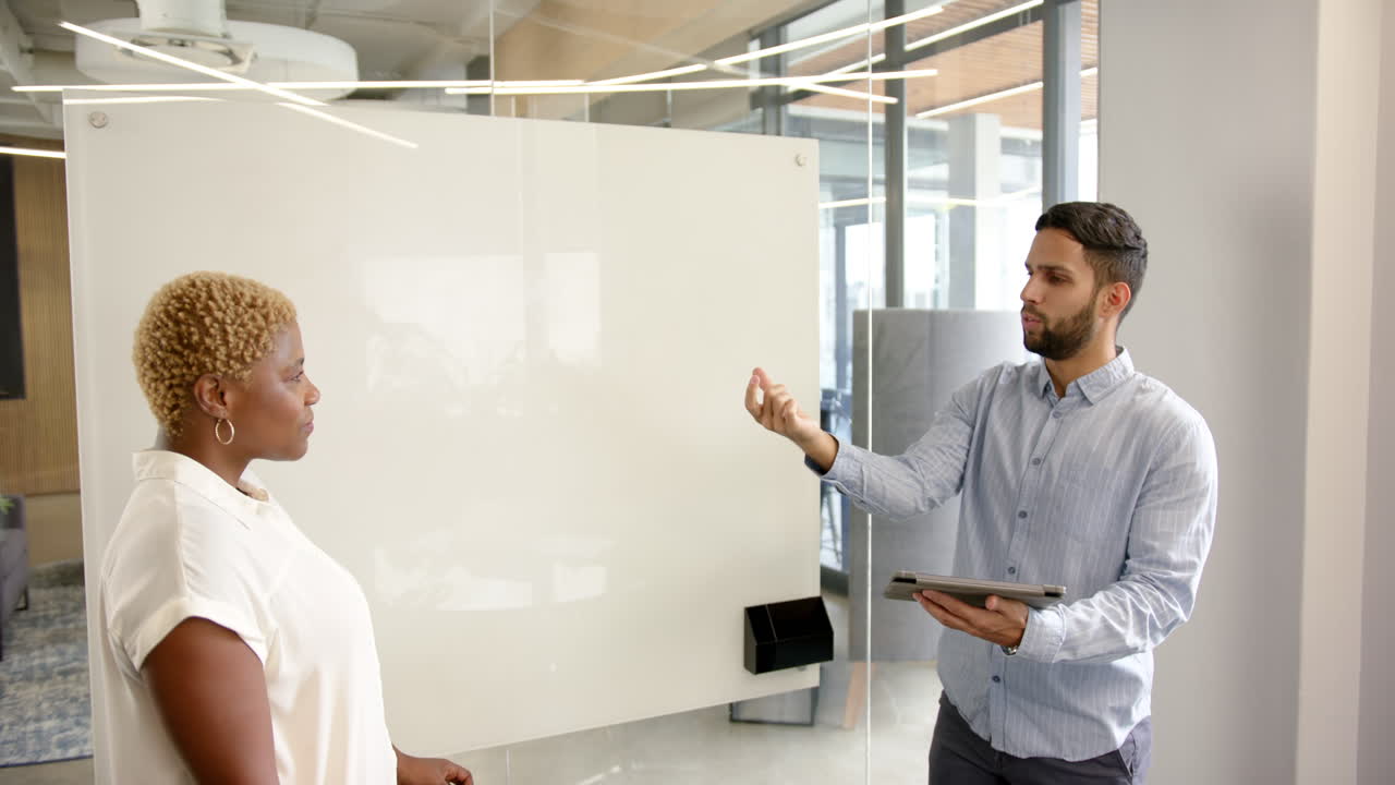 Discussing project details, diverse colleagues using tablet and whiteboard in modern office