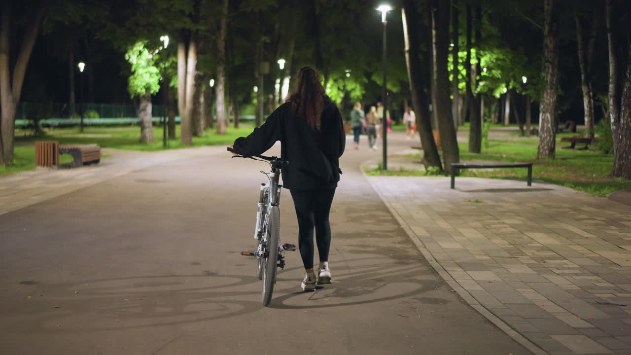 Night Park Approach Caucasian Woman Bicycle, Walking Forward Toward Distant Pedestrians And Benches, Soft Lamplight, Casual Hoodie, Urban Pathway With Trees And Ambient Glow, Curious Mood