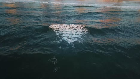 Toma-Aérea-De-Drones-Durante-El-Amanecer-Sobre-Olas-Con-Gente-Caminando-En-La-Playa