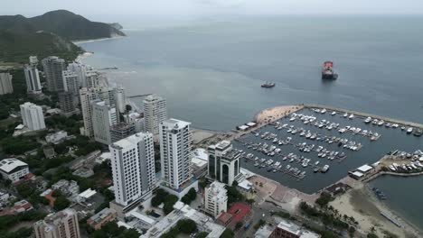 Flying-Above-Santa-Marta-City-Marina,-Colombia,-Aerial-View-of-Sailboats,-Breakwater-and-Cityscape-Skyline