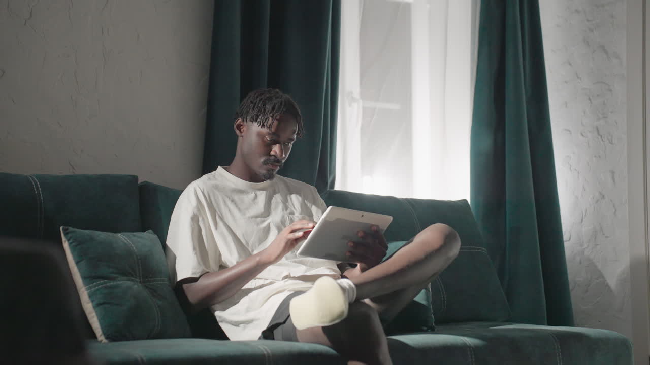 Champ focused on tablet, seated on teal sofa in bright living room. Sunlight shines through window, creating a calm and cozy space for work or relaxation. Peaceful, modern atmosphere