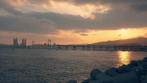 view of gwangan bridge with dramatic sky during golden sunset in busan, sun setting over mountain with sunlight reflections in sea waters