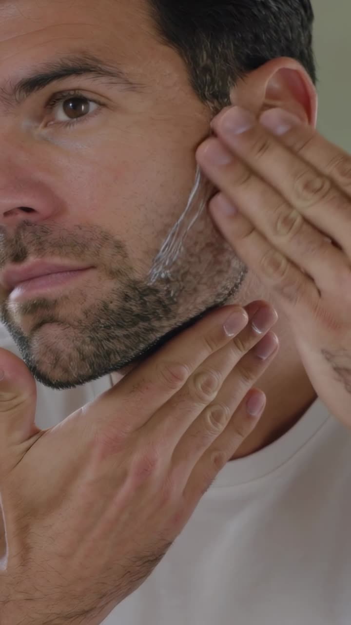 Video verticale: Uomo che ispeziona la mascella allo specchio, controlla la barba incolta davanti allo specchietto, applicando il gel da barba
