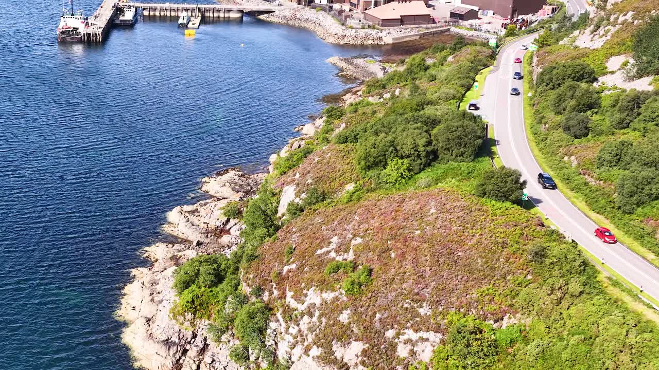 Drone glides over winding coastal road toward harbor, boats, and village under bright daylight